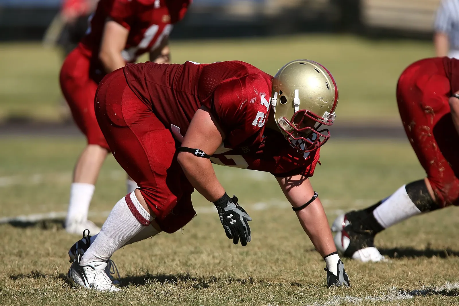 A lineman in a red jersey and gold helmet crouched low in a three-point stance at the line of scrimmage, other red-jerseyed teammates blurred behind him.