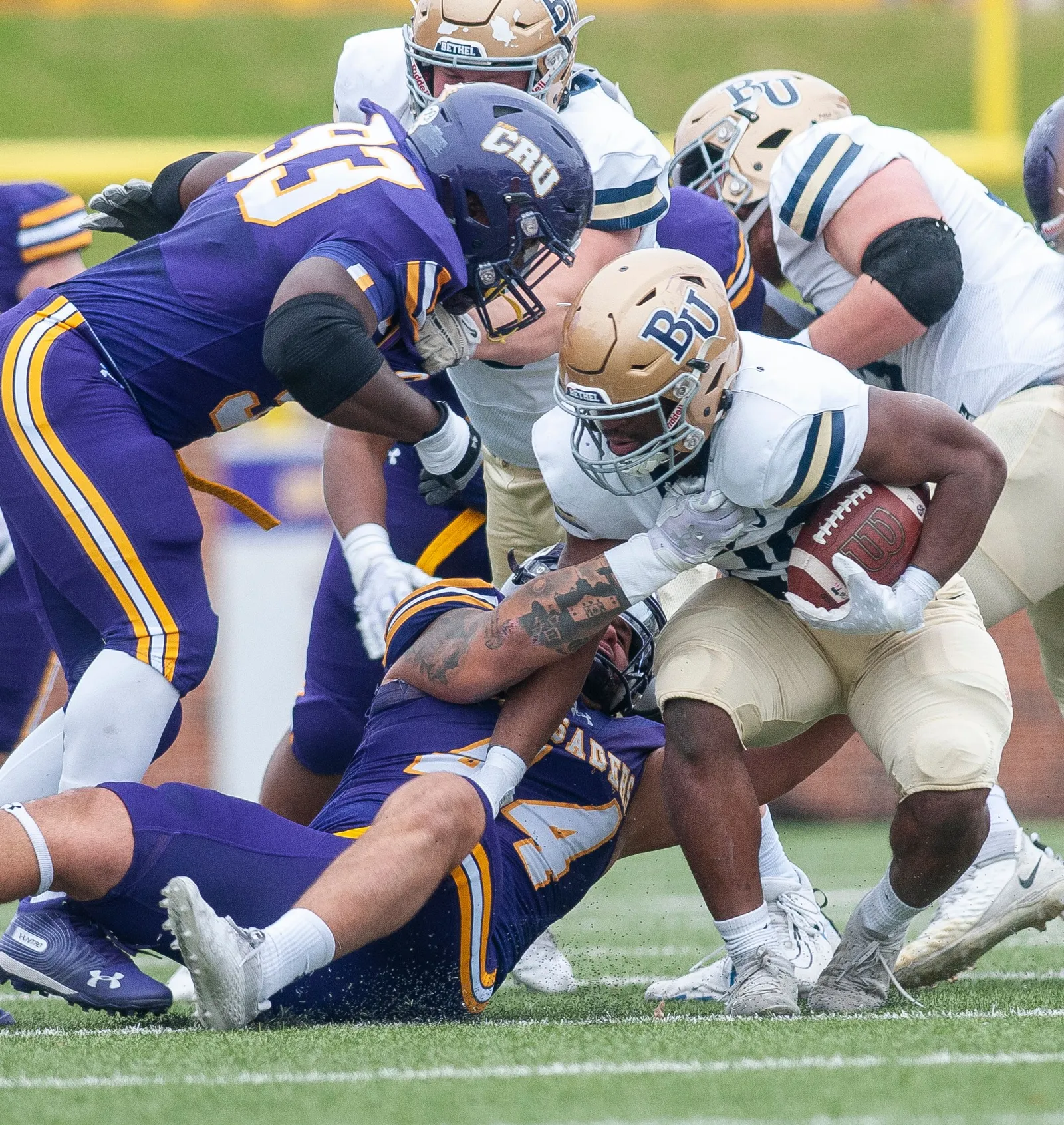 A ball carrier in a white and gold uniform is gang-tackled by defenders in purple as the play is stopped near the line of scrimmage.