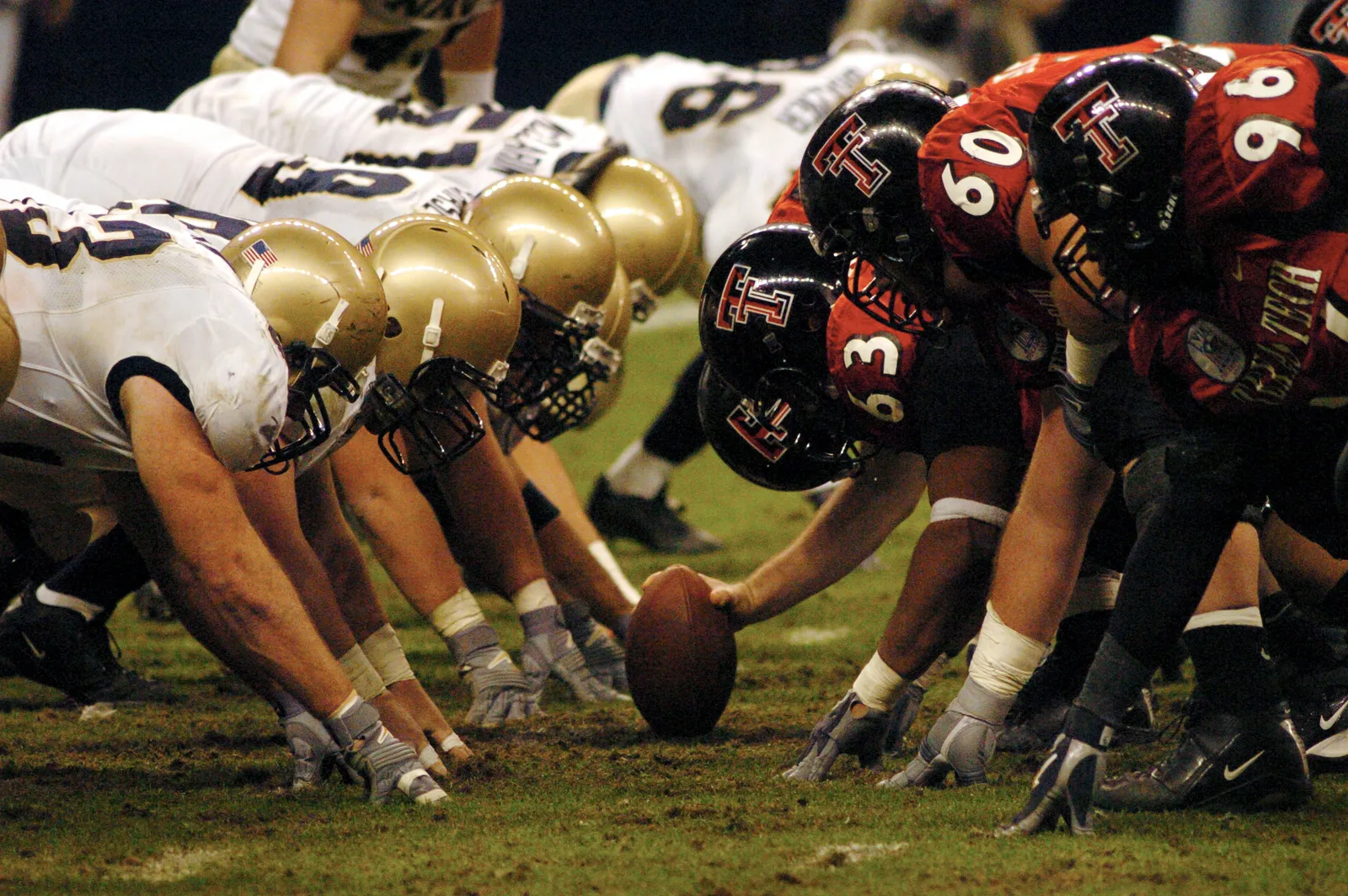 A close, low-angle view of two college football lines squared off nose-to-nose at the line of scrimmage in the seconds before the snap, one team in black helmets with red jerseys, the other in gold helmets with white jerseys.