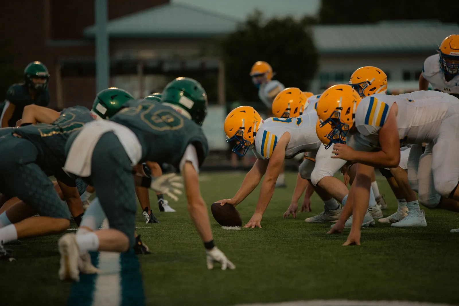 High school offensive and defensive linemen set against each other at the line of scrimmage under stadium lights, one player's hand on the football an instant before the snap.