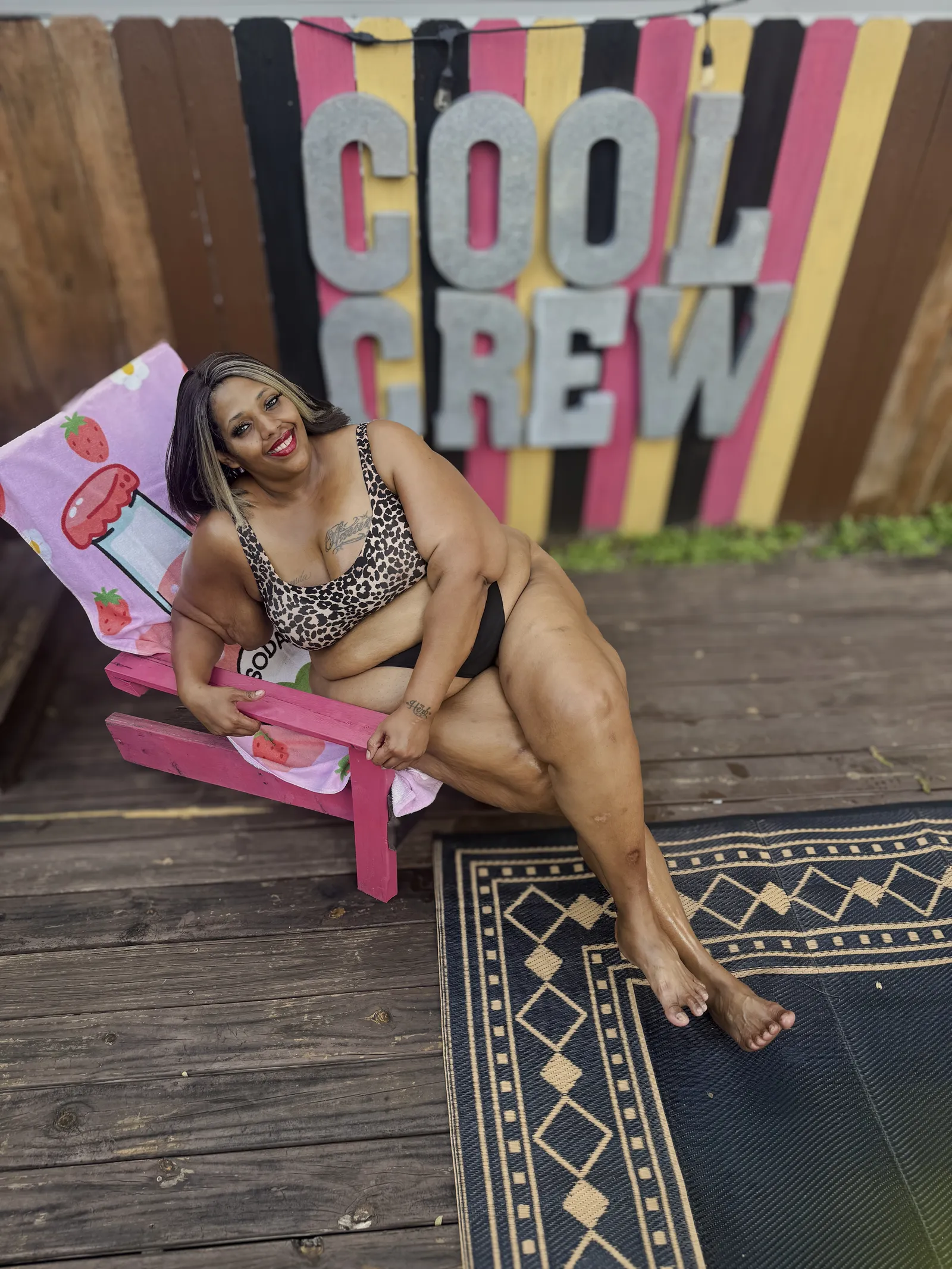 Amanda seated sideways across a pink wooden deck chair with a strawberry-print towel over it, smiling toward the camera, in front of a fence painted in pink, black and yellow stripes with large metal letters reading COOL CREW.