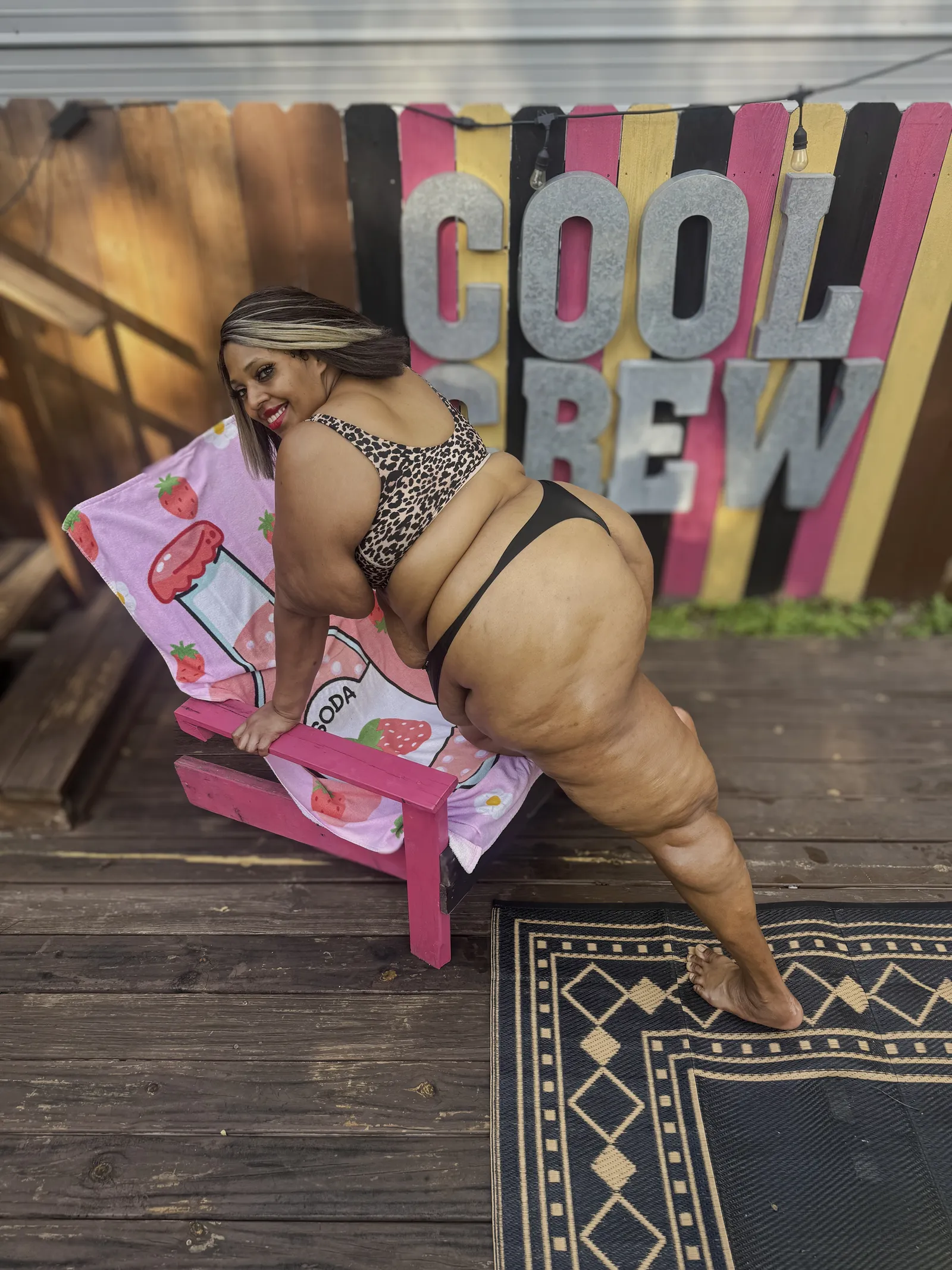 Amanda bracing on the pink deck chair and looking back over her shoulder, the metal COOL CREW letters and a string of bulbs on the fence behind her.