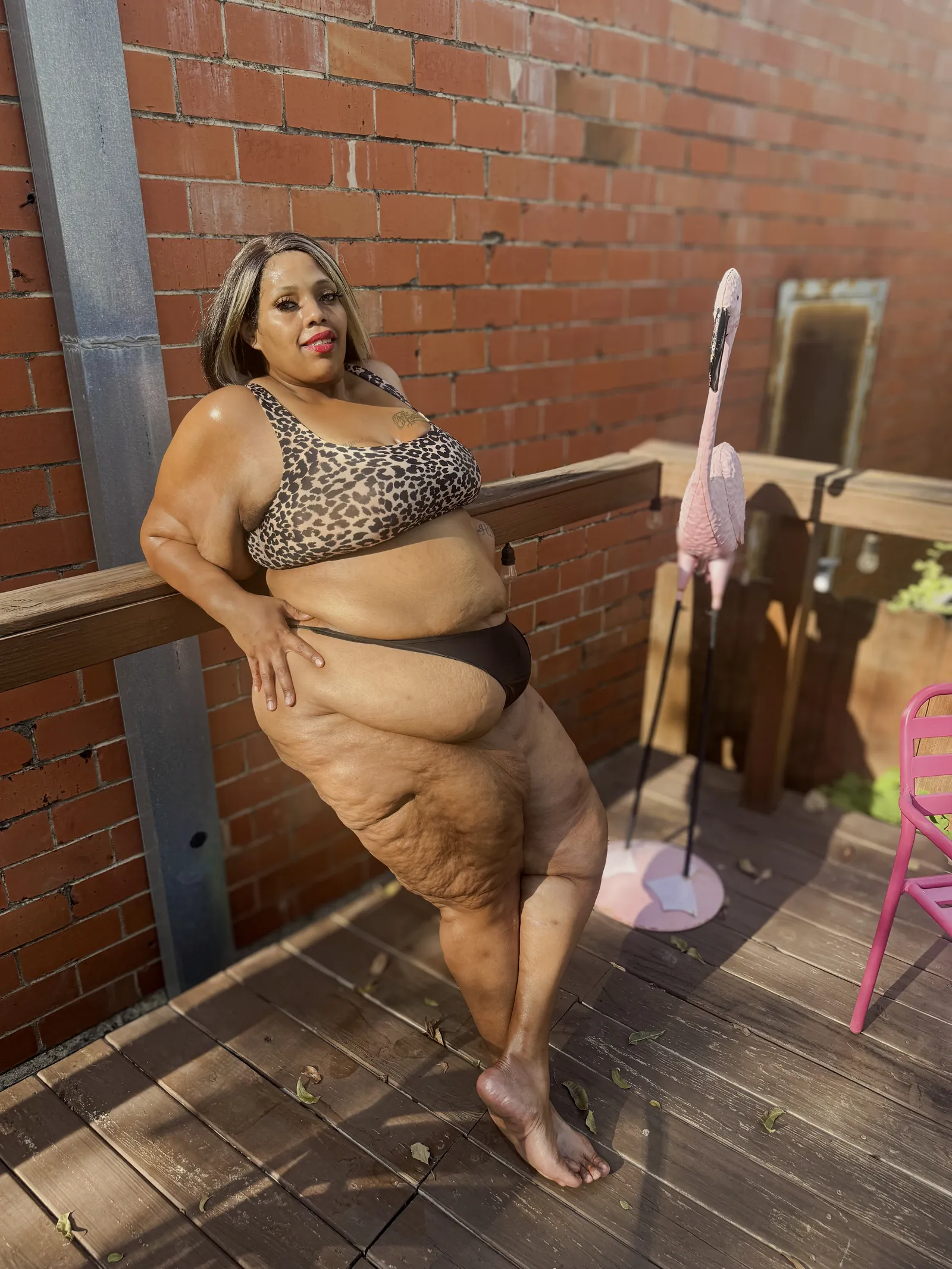 Amanda standing against a wooden deck rail in front of a red brick wall, one hand resting on her hip, a pink flamingo lawn ornament and a pink metal chair beside her.