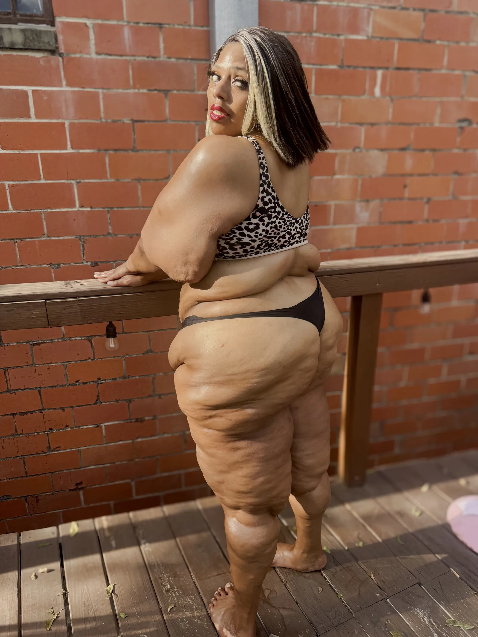 Amanda resting a forearm on the deck rail and looking back over her shoulder at the camera, red brick wall behind her.