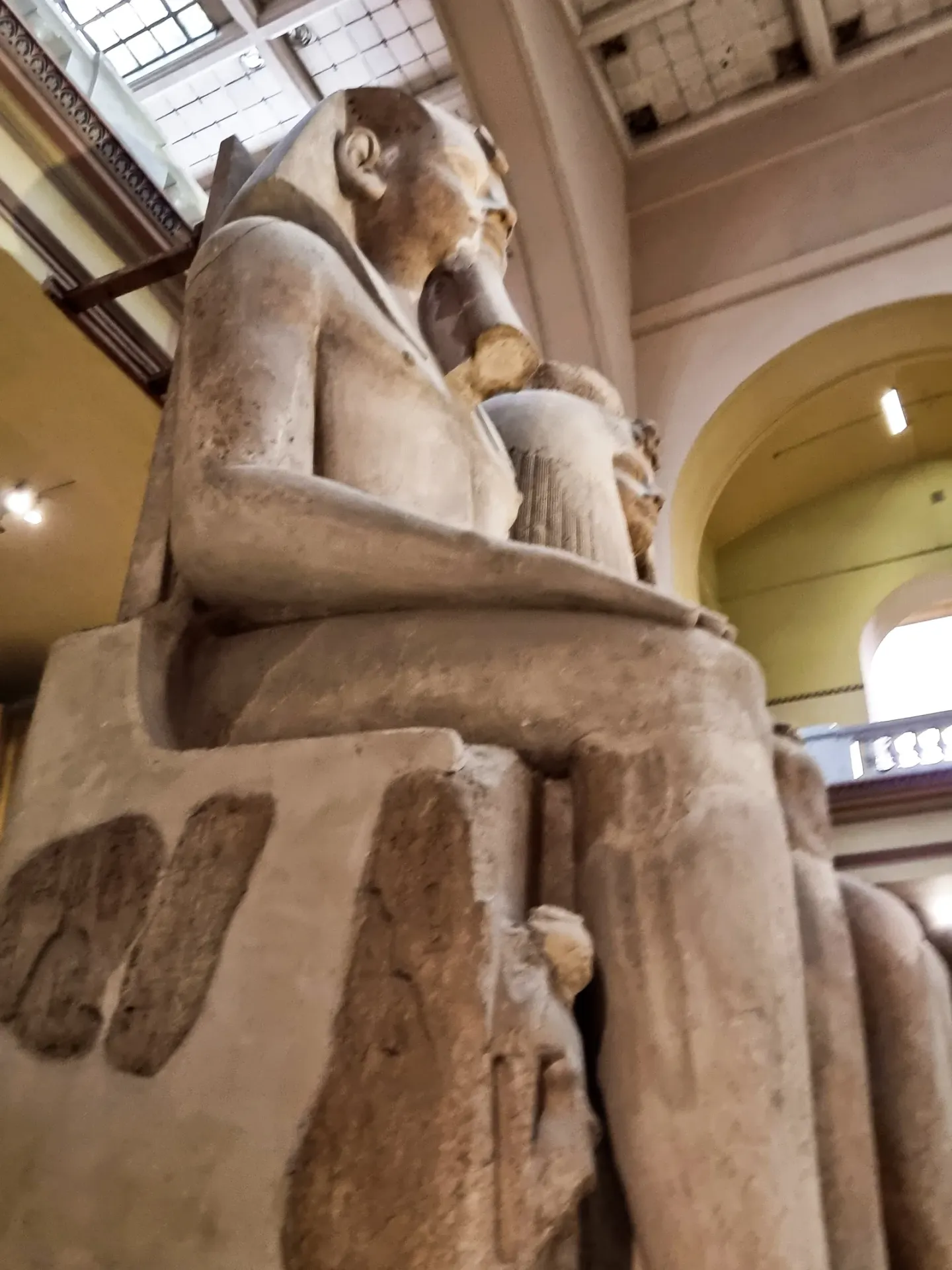 A colossal ancient Egyptian statue of a seated pharaoh photographed from below in a museum hall, its hands resting flat on its knees, daylight falling through the arched windows behind it.