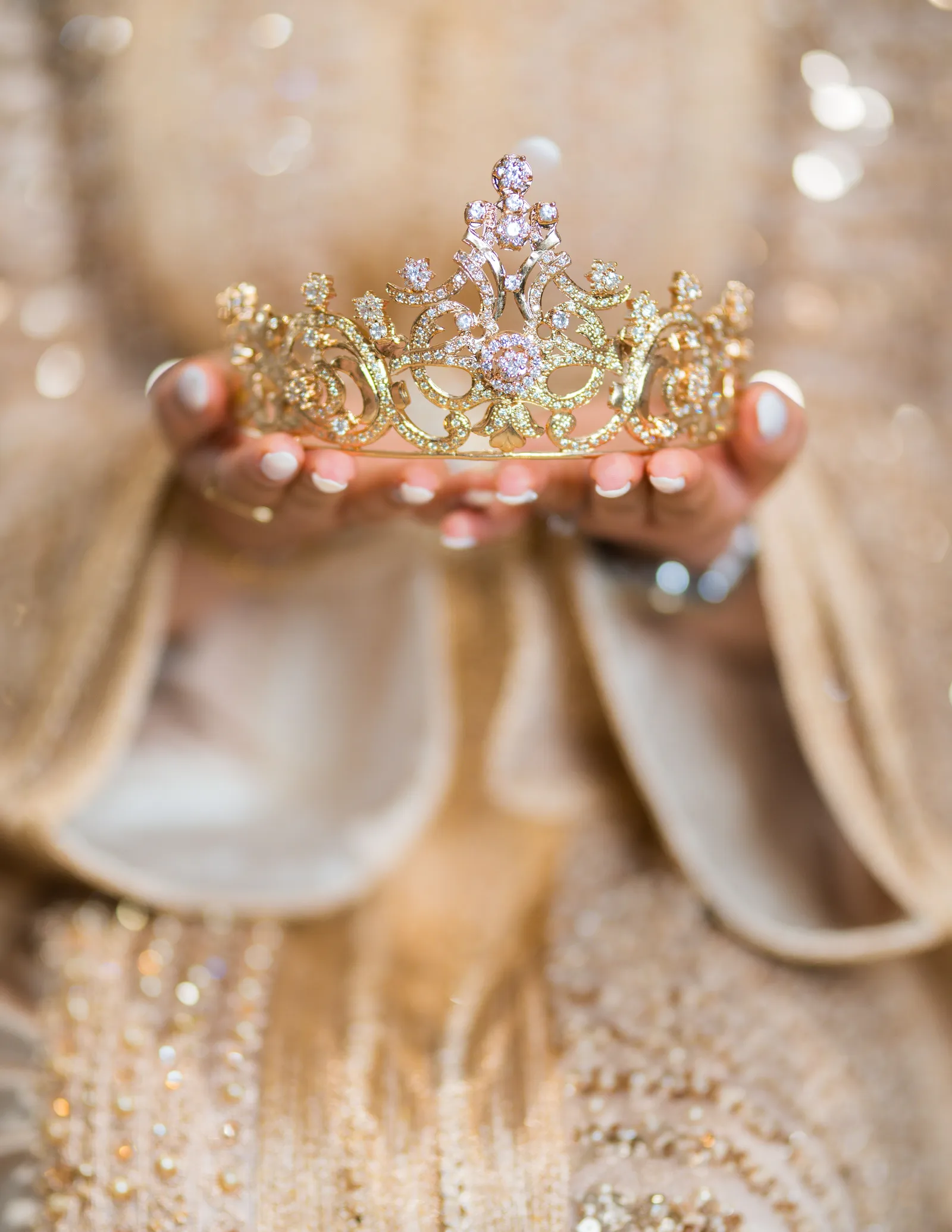 A jeweled gold tiara held delicately in a pair of outstretched, unidentifiable hands. No face is visible and no specific person is identifiable.
