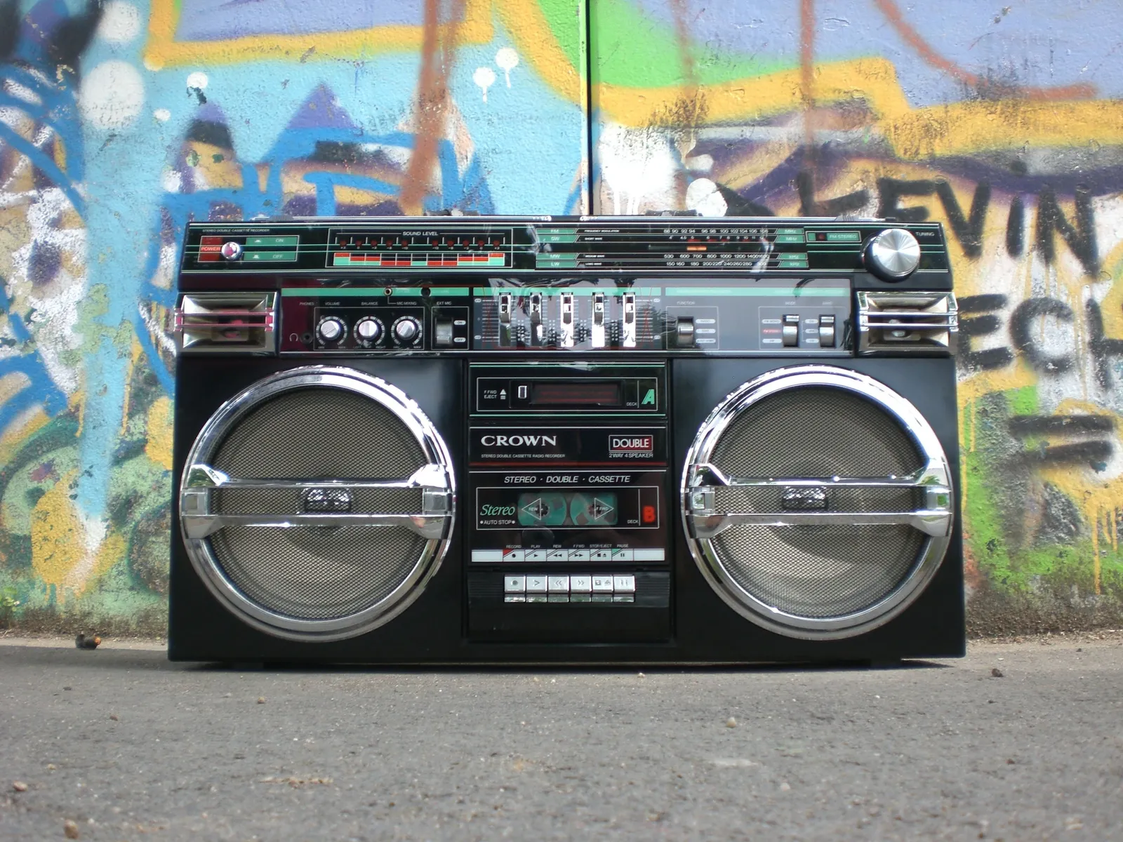 A vintage boombox with twin speakers and a double-cassette deck, set on the ground in front of a graffiti-covered wall. No people are depicted.