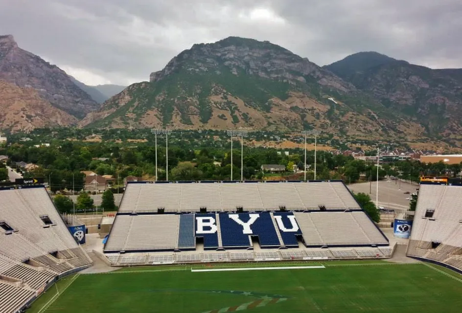 An elevated view of LaVell Edwards Stadium's empty seating bowl, with white and navy seats spelling out "BYU" and the Wasatch Range rising behind the stadium.