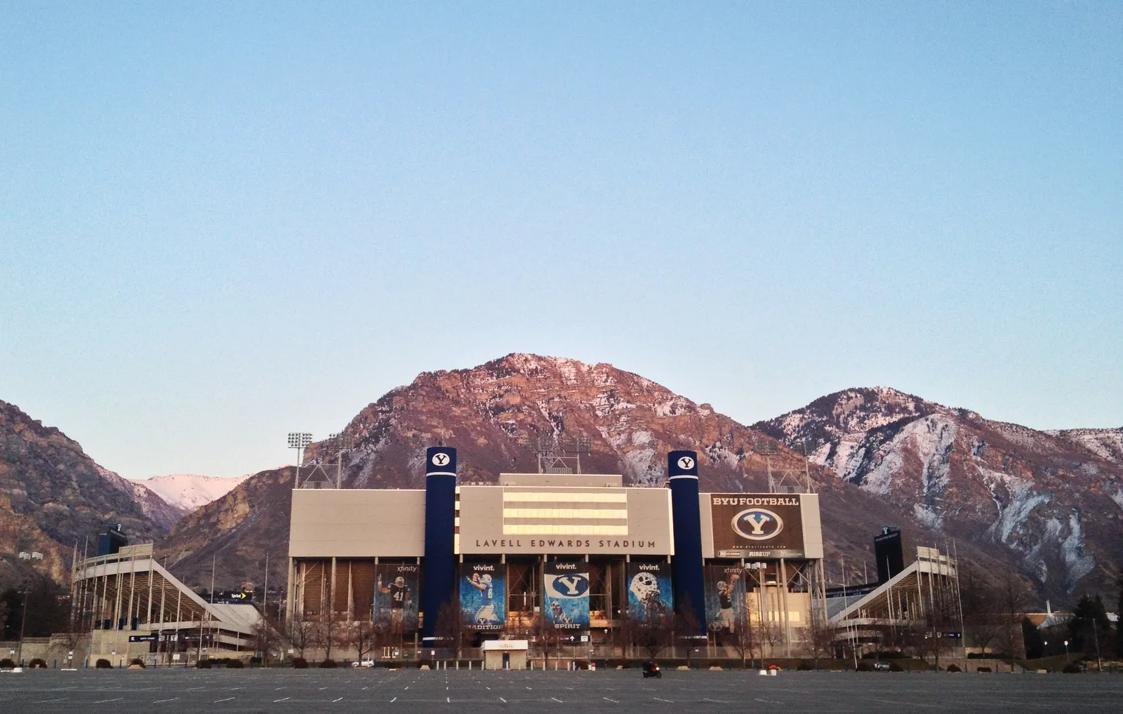 LaVell Edwards Stadium's main entrance lit at dusk, with the Wasatch mountains rising behind it and an empty parking lot in the foreground.