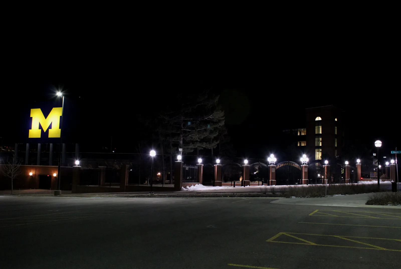 The gated main entrance to Michigan Stadium at night, with the University of Michigan's illuminated block-M sign at left and "Michigan Stadium" lettering on the brick gate — the actual venue that hosted Saturday's Oklahoma game.