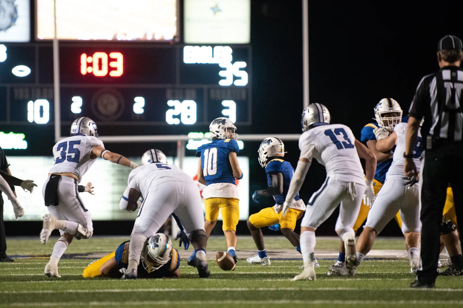 Two football teams in blue-and-white and white uniforms scramble for a loose ball on a lit stadium field at night, a referee and scoreboard visible behind them. No Michigan or Oklahoma team markings are shown.