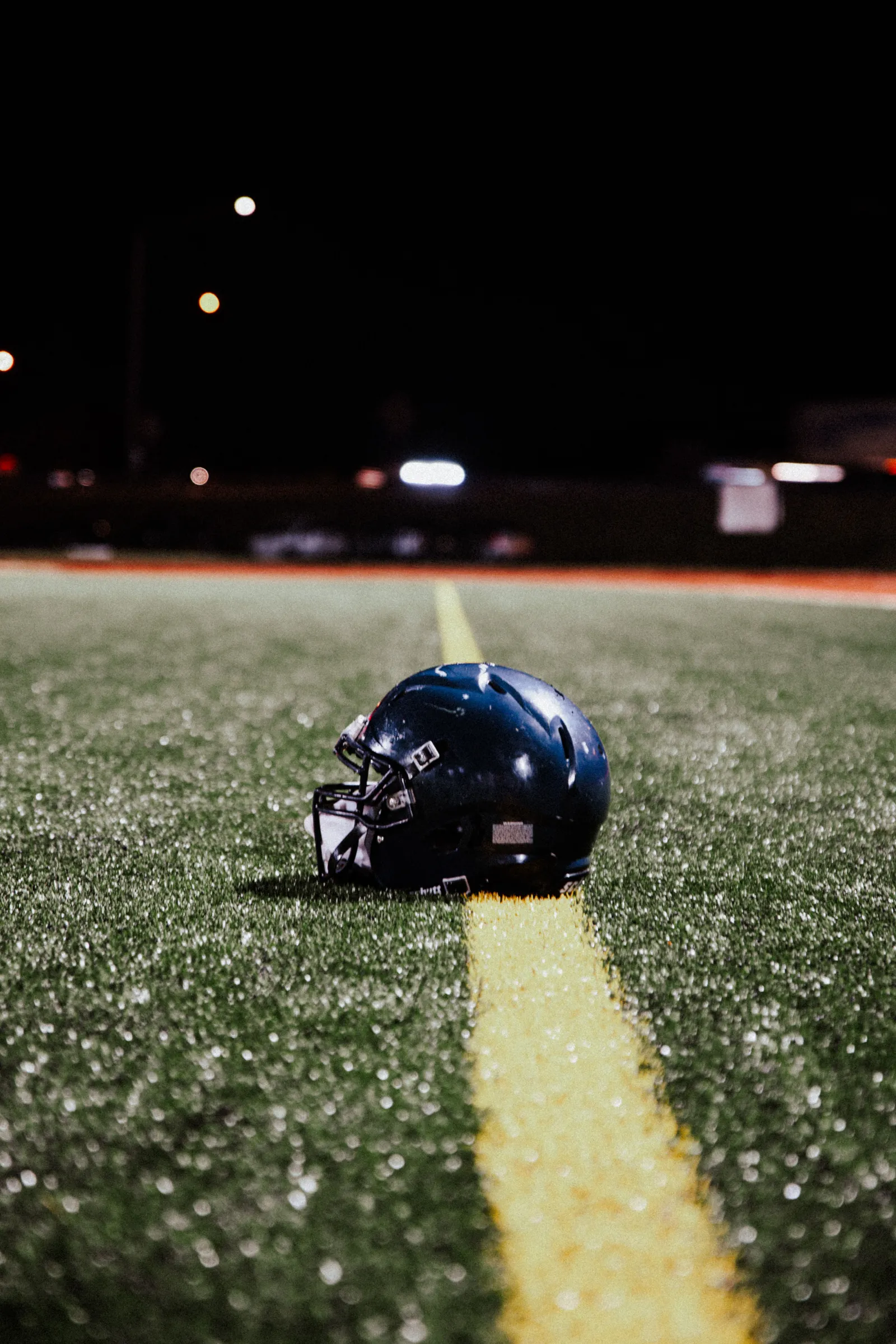 A single dark football helmet resting on turf beside a yellow field line at night, stadium lights and blurred parked cars visible in the background — no team markings visible, an unrelated venue used here as a neutral pre-kickoff image.