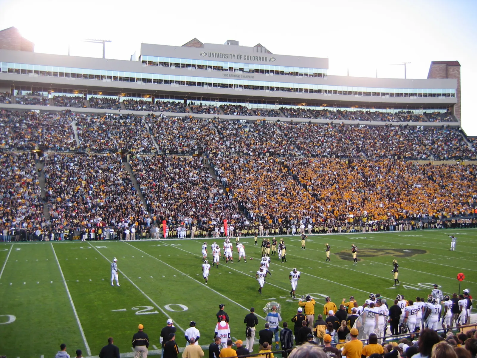 A packed Folsom Field crowd in black and gold watches a Colorado Buffaloes home game, with players from both teams lined up on the field below.