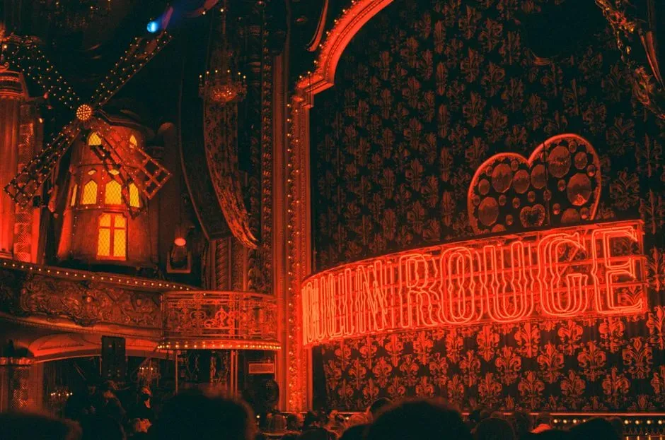 The facade of a theatre at night, its red neon signage and illuminated windmill glowing above the silhouetted heads of a crowd waiting outside.