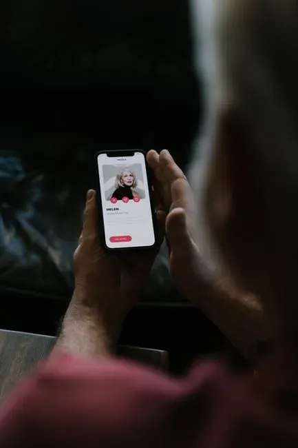 A person seen from behind in a dark room holding up a smartphone, a dating profile card with a photograph and a red action button filling the screen.