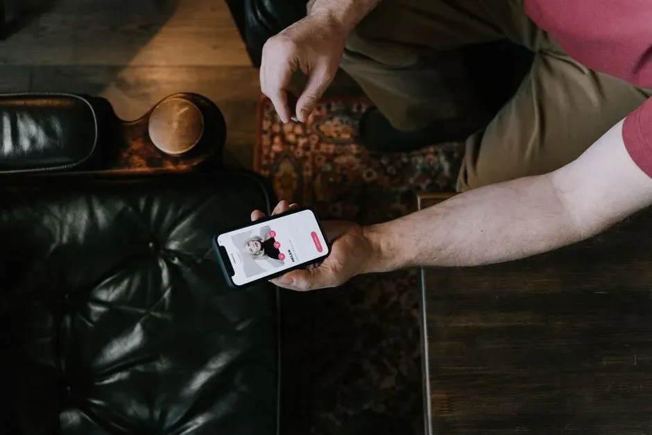An overhead view of someone sitting on a dark leather sofa holding a phone showing a dating profile, a bowl of snacks and a mug on the table beside them.
