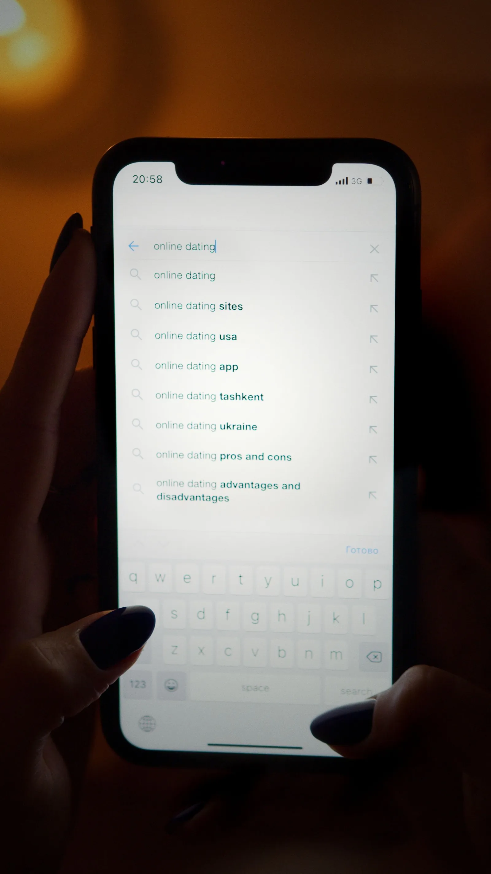 A hand holding a smartphone at night, the screen showing a list of autocomplete search suggestions for online dating above an open on-screen keyboard.