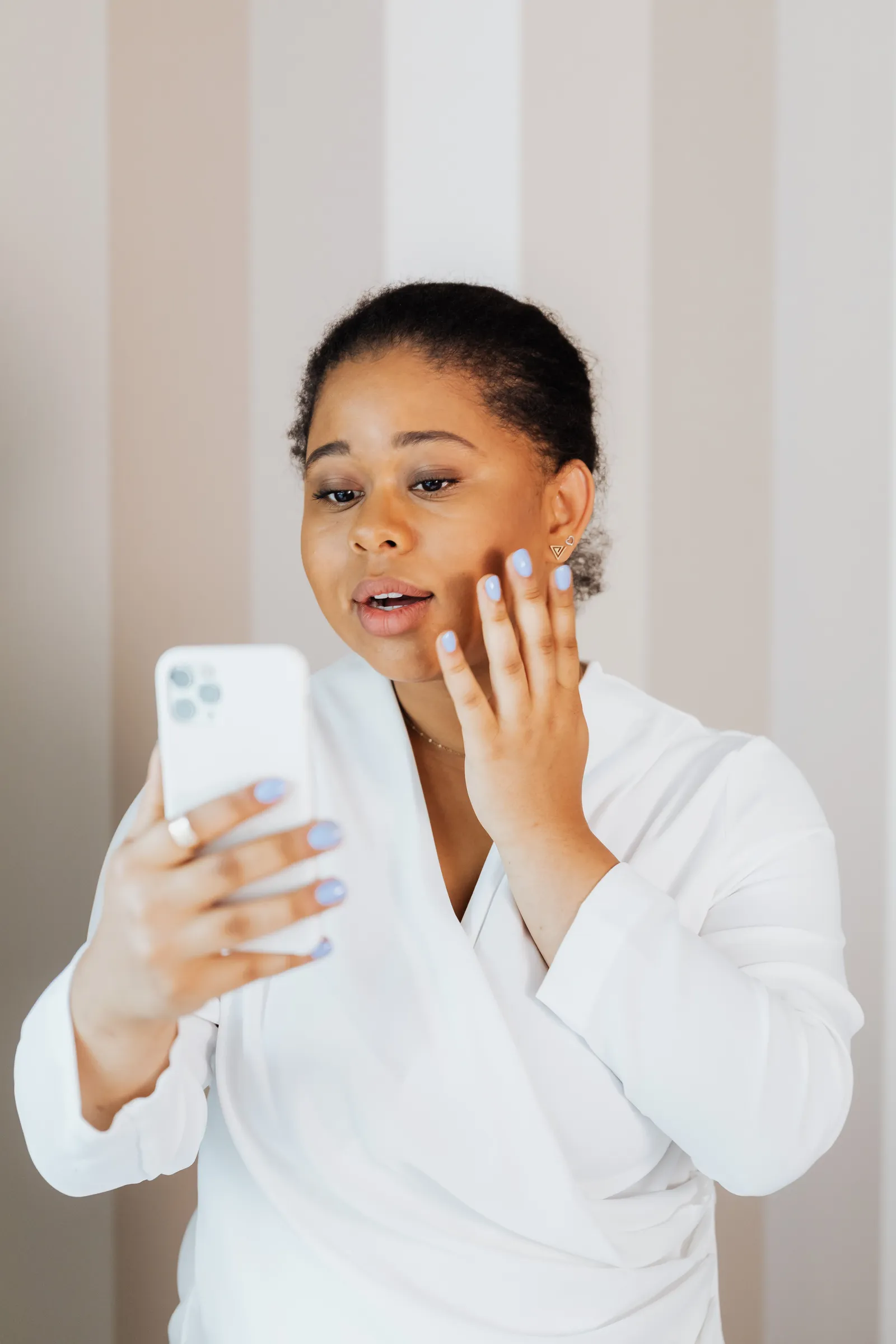 A woman in a white top holding up her smartphone and touching her own cheek as she studies her face on the screen — the gesture of lining up a verification selfie.