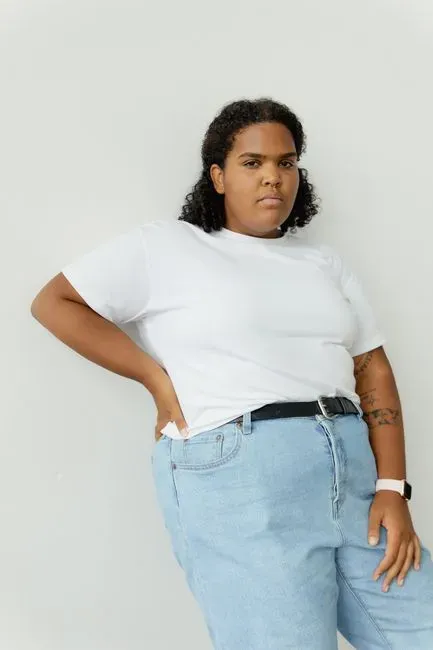 A plus-size woman standing against a plain pale backdrop in a white T-shirt tucked into light blue jeans with a black belt, one hand in her pocket, the waistband sitting flat against her body.