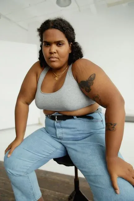 The same model perched on a stool in light blue jeans and a grey ribbed crop top, leaning forward with her hands on her thighs so the fit across the hip and thigh is visible.