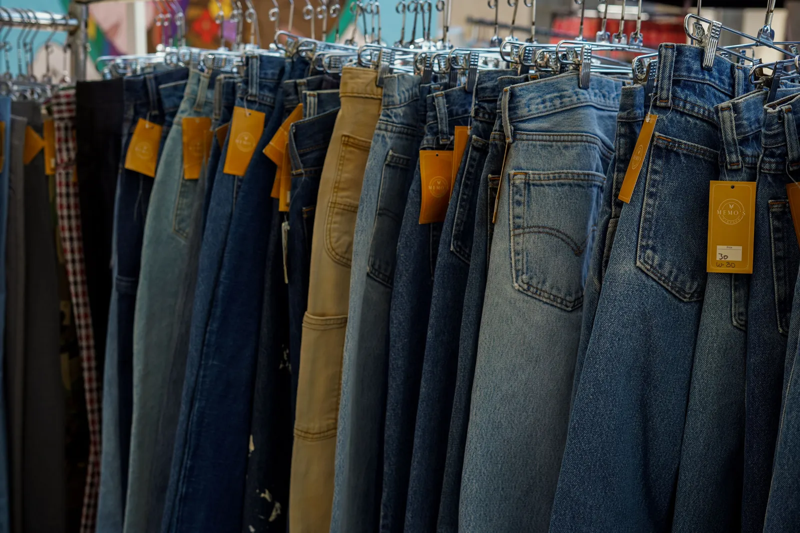A dense rail of jeans on metal hangers in a store, price tags looped through the belt loops, the denim ranging from pale stonewash to deep indigo.