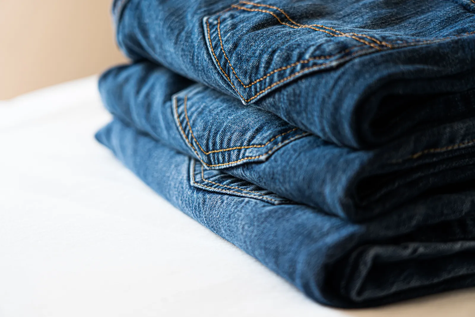 A neat stack of three pairs of folded blue jeans on a white surface, photographed close so the topstitching and thick waistband seams are clearly visible.