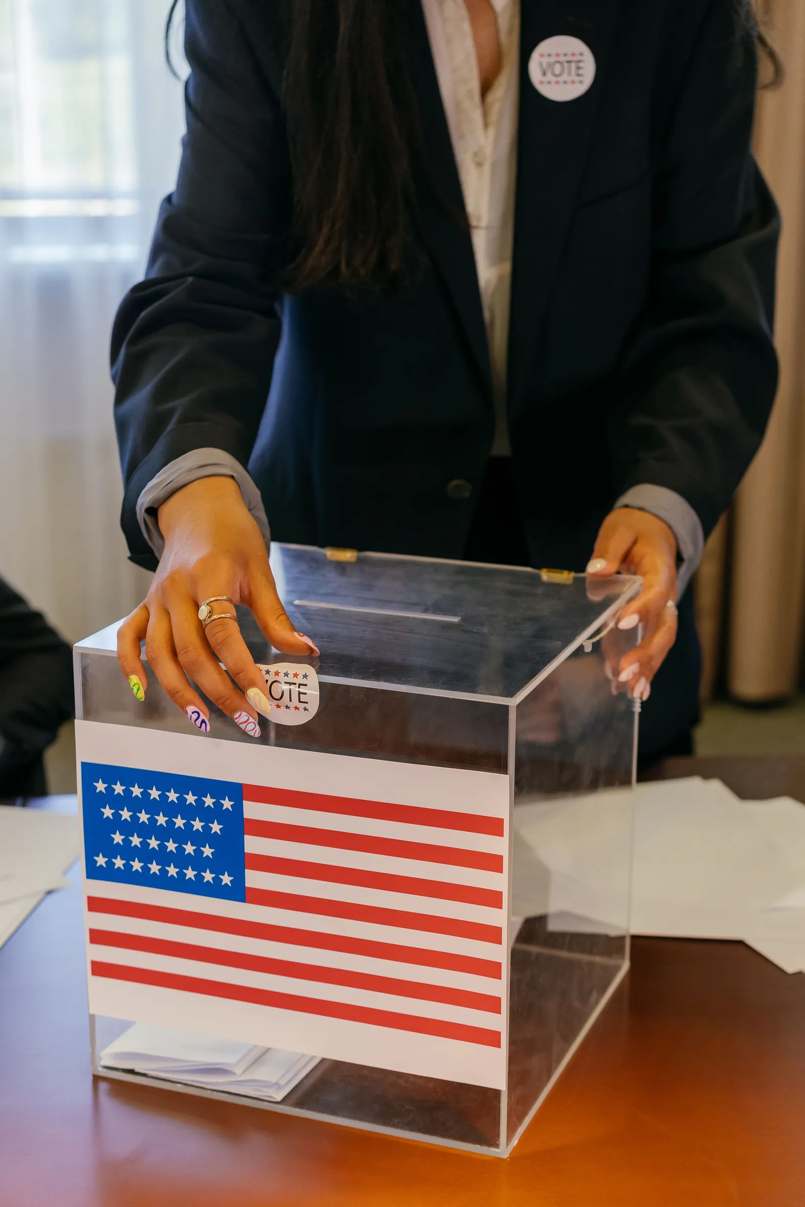 Close-up of a person casting a vote into a transparent ballot box beside an American flag; the voter's face is not visible.