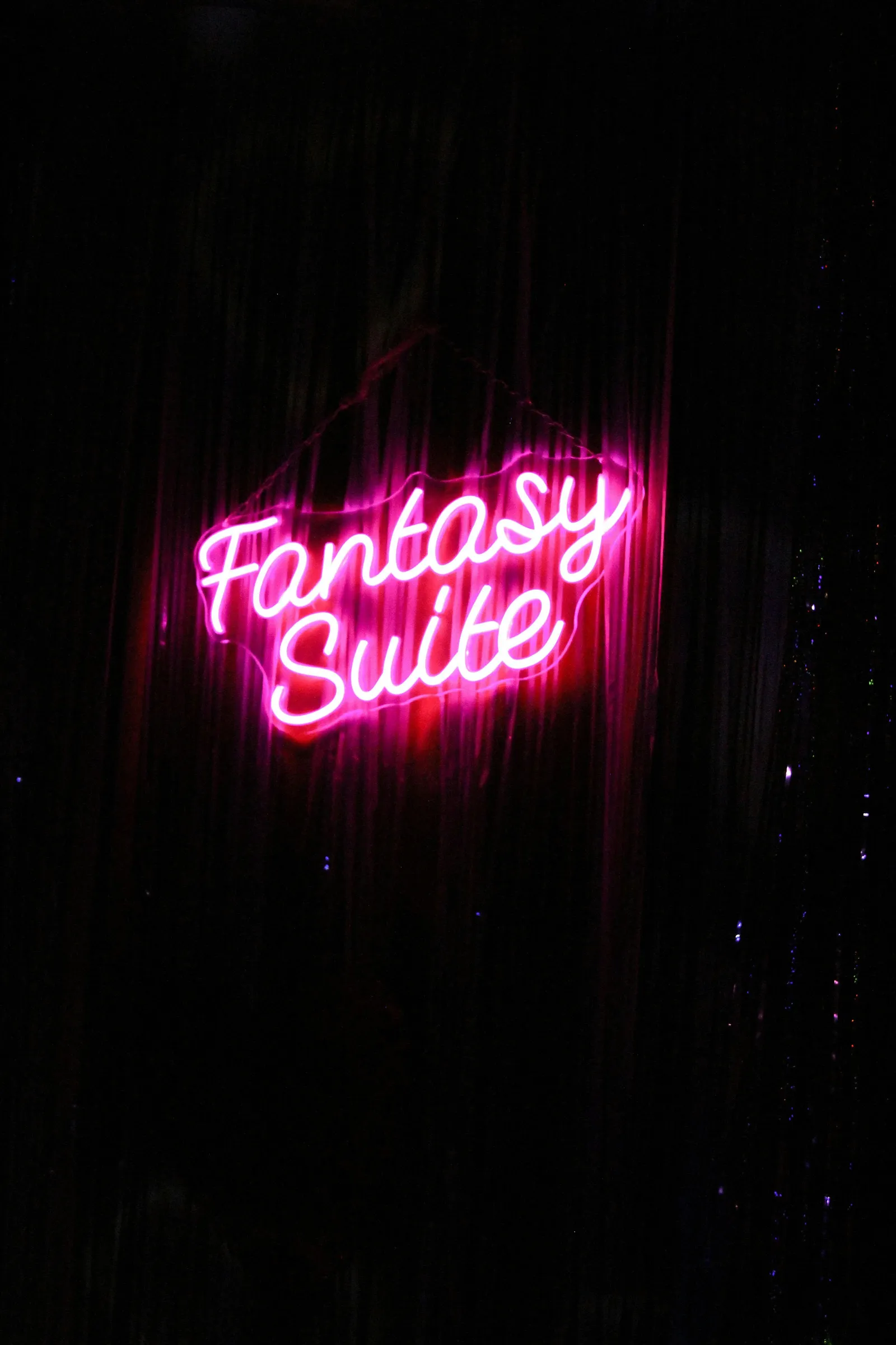 A photograph of a pink neon sign reading Fantasy Suite in cursive script against a dark wall.