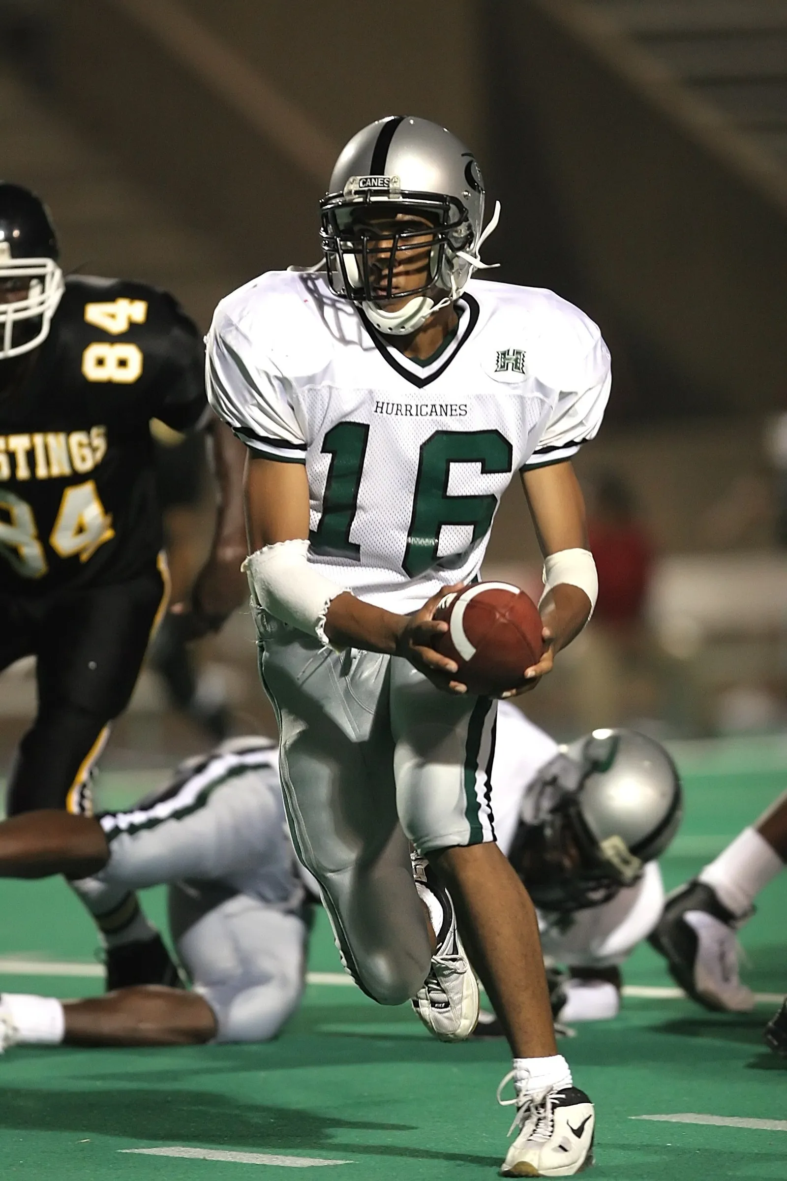 A high school football player in a white and green 'Hurricanes' uniform (No. 16) carries the ball upfield with both hands during a game, with a black-jerseyed defender in pursuit and other players on the ground nearby. No NFL team markings are visible.