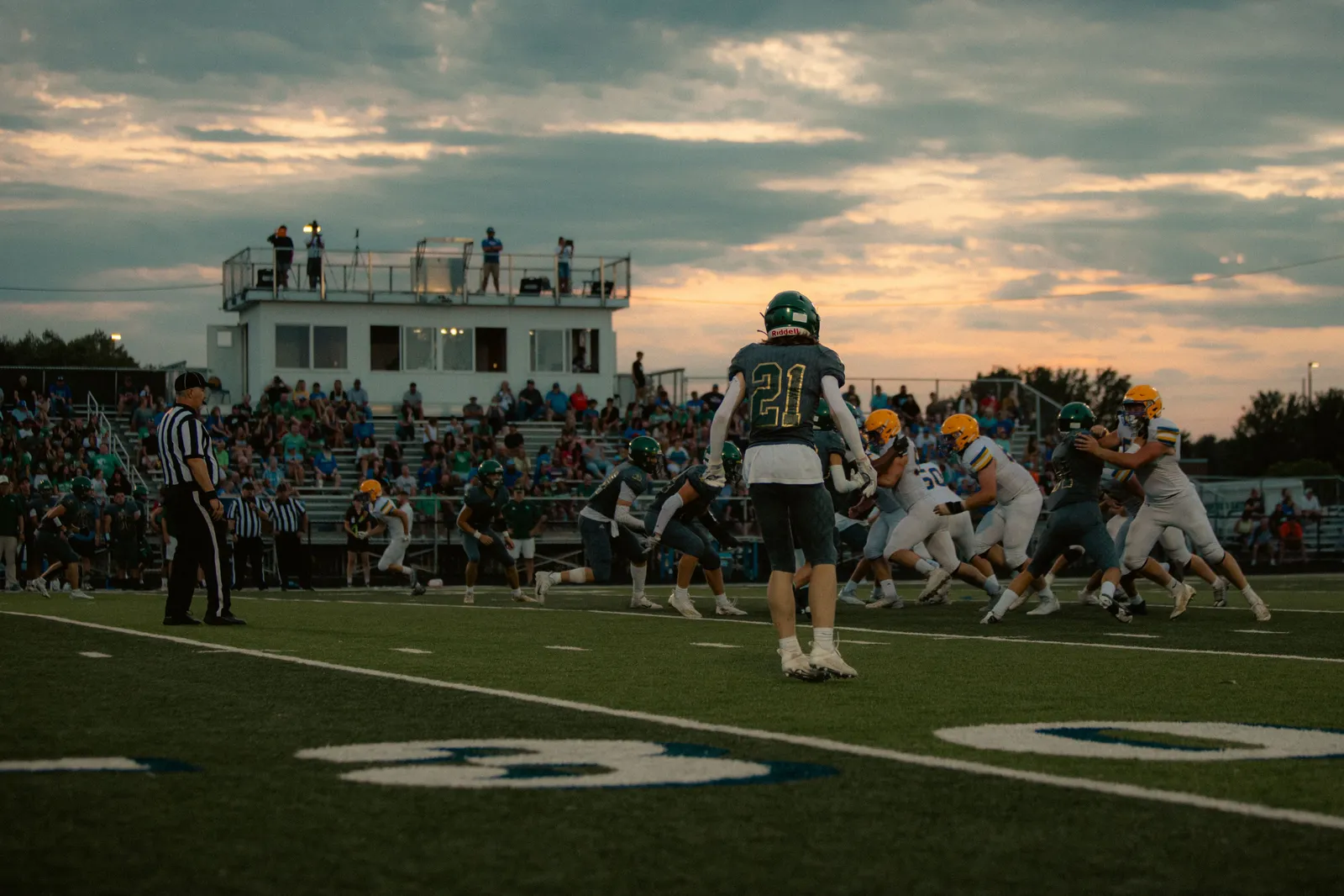 A high school football game at dusk: a defensive player in a dark green jersey (No. 21) walks toward the line of scrimmage as a referee looks on and a packed crowd fills the bleachers behind the field. No NFL team markings are visible.