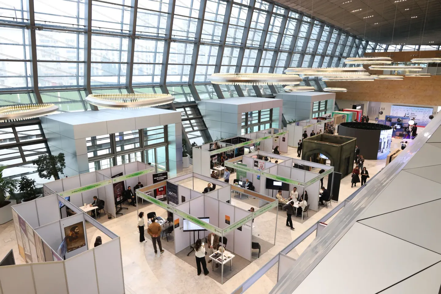 A large glass-roofed exhibition hall seen from a balcony, filled with rows of white modular trade-show booths and small groups of visitors moving between them.