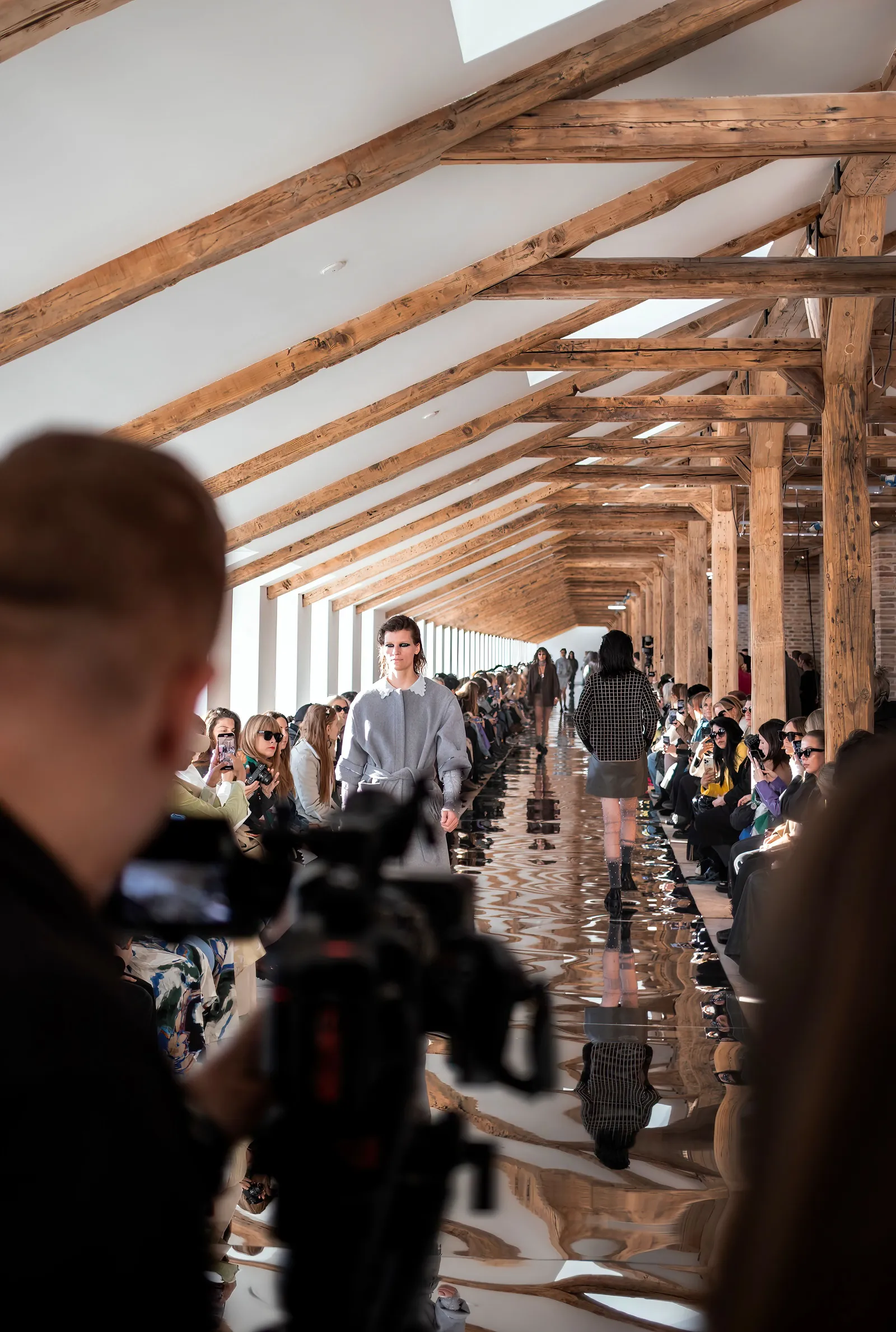 A model walking a runway down the centre of a long timber-beamed hall, seated guests lining both sides and a photographer shooting from the end of the catwalk in the foreground.