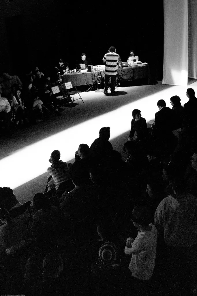 A black-and-white photograph of a fashion show in progress, a single model lit on a bright runway with the silhouetted heads of the seated audience filling the dark foreground.