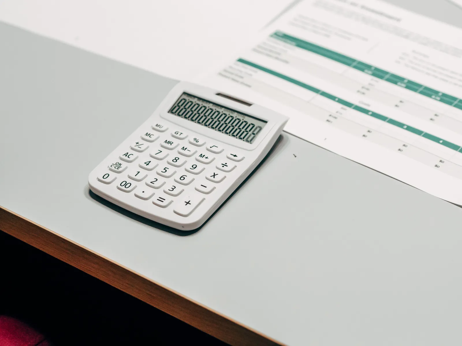A small white desk calculator showing a row of zeros on a pale desk, beside a printed sheet of figures ruled into green and teal shaded columns.