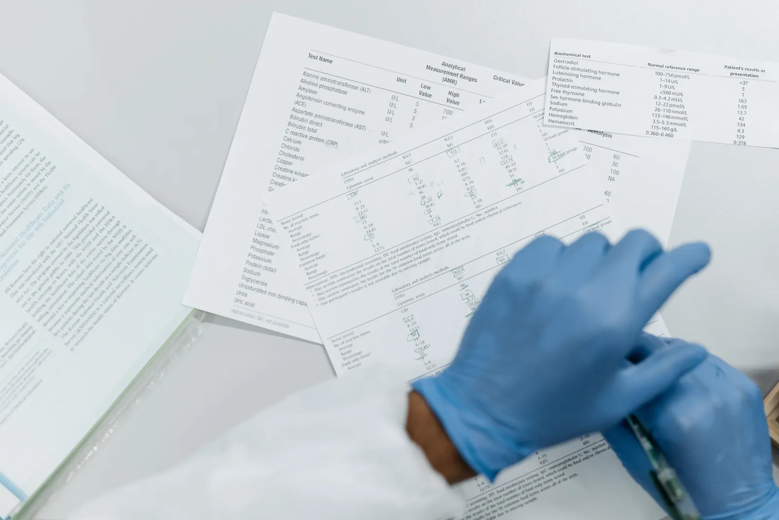 Blue-gloved hands resting over overlapping printed pages of laboratory reference ranges and test results spread across a white bench, an open binder at the left edge.