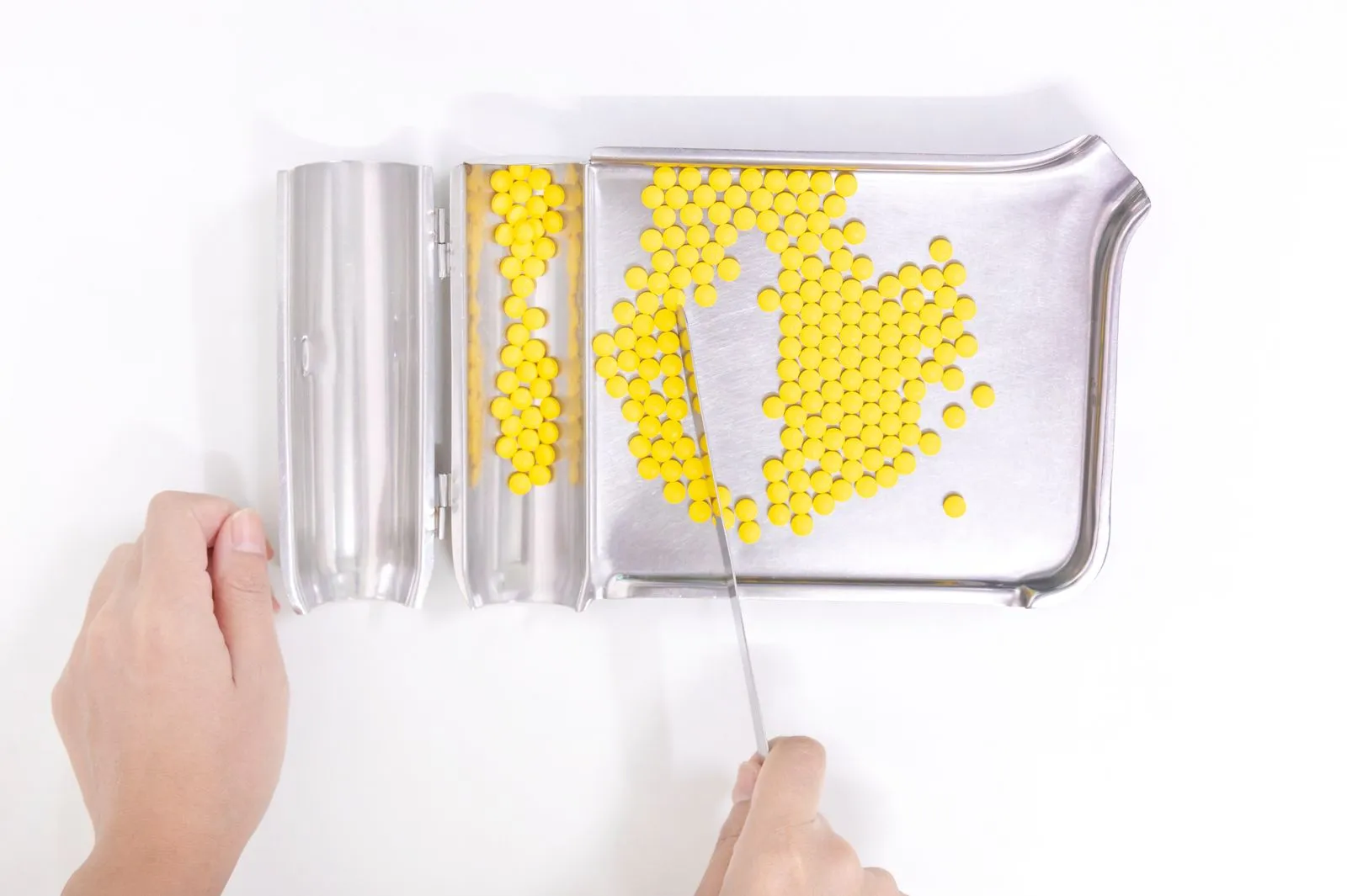 A pharmacy technician's hands counting small yellow tablets on a stainless steel dispensing tray with a metal spatula, seen from above on a white counter.