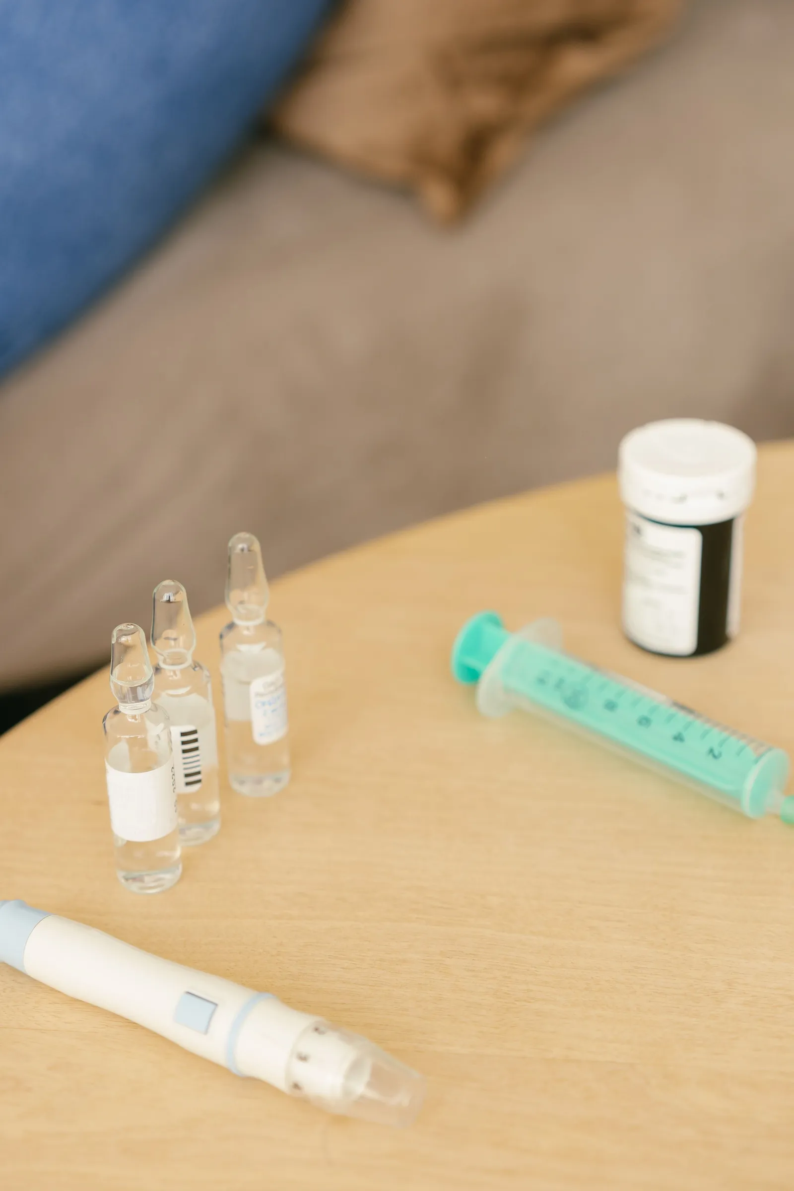 Small glass medication vials, a green plastic syringe, a white pill bottle, and an injection pen arranged on a wooden side table.