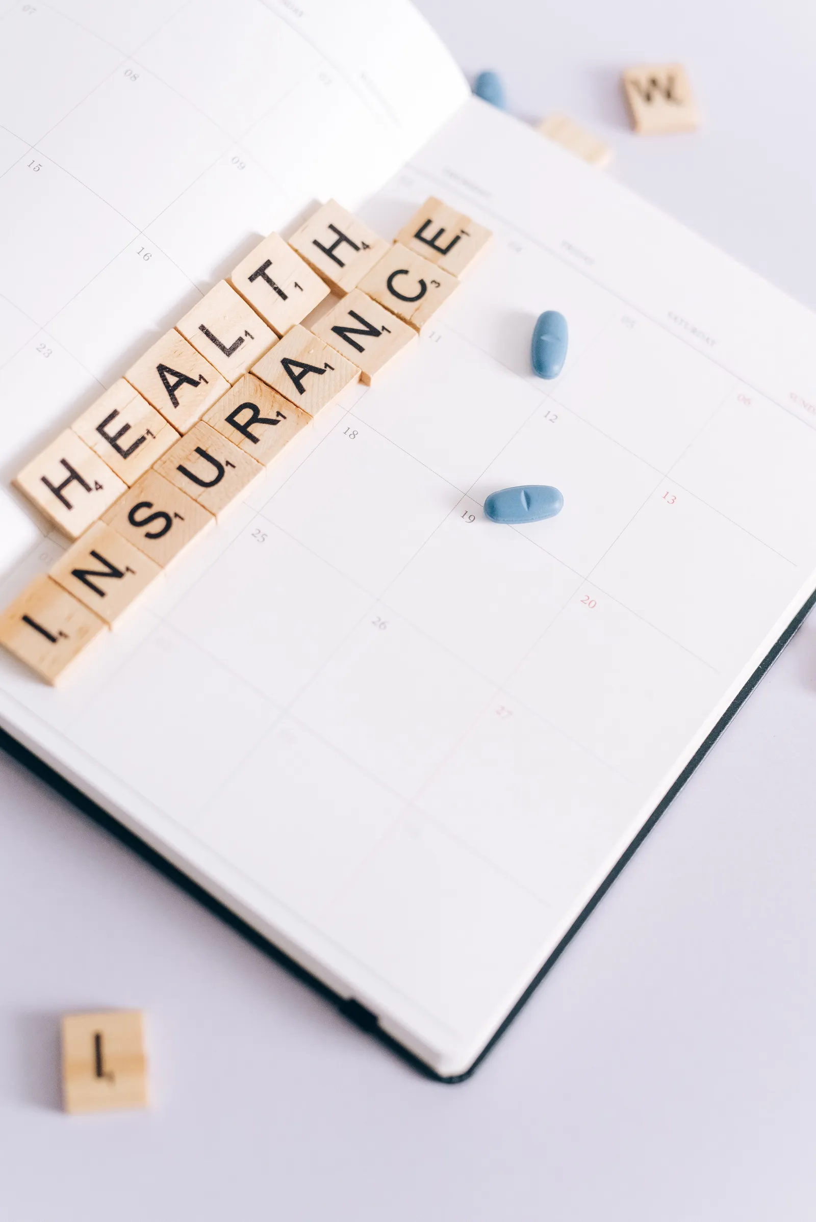 Scrabble tiles spelling out 'health insurance' arranged on a printed calendar page beside a small pile of blue pills.