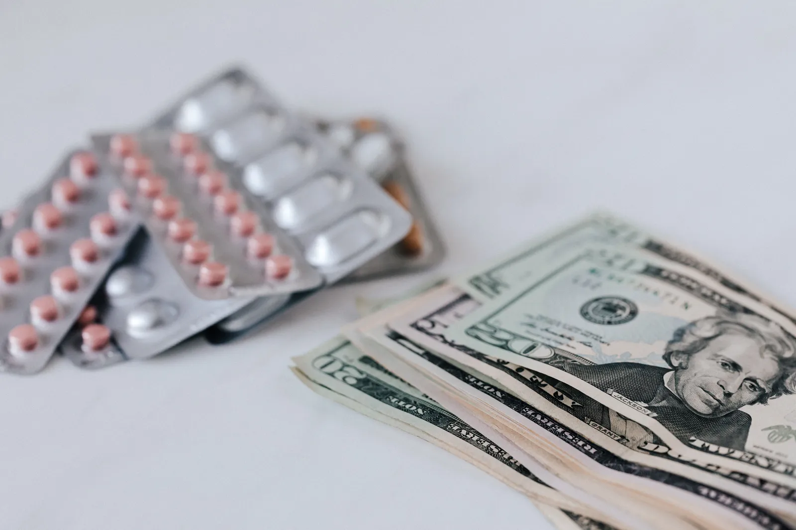 A loose pile of American dollar bills next to blister packs of medication on a white table.