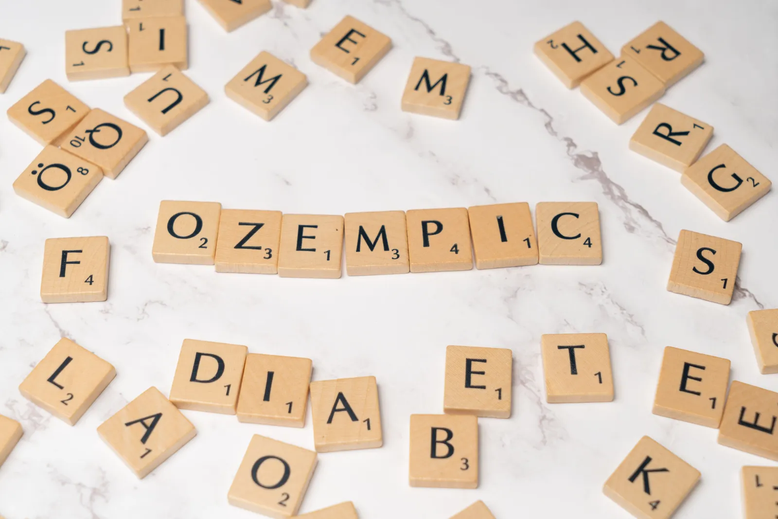 Wooden Scrabble tiles spelling 'Ozempic' and 'diabetes' scattered across a white marble surface.