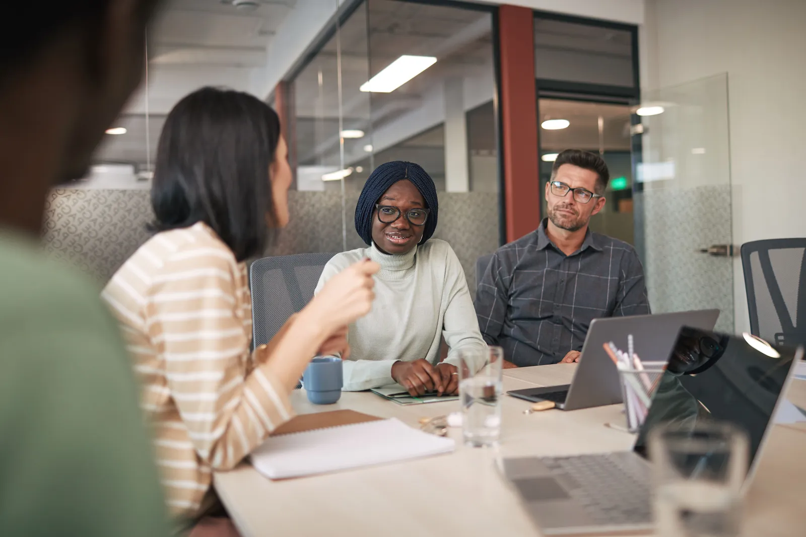 Four professionals in business attire around a conference table during an office meeting, laptops open in front of them.