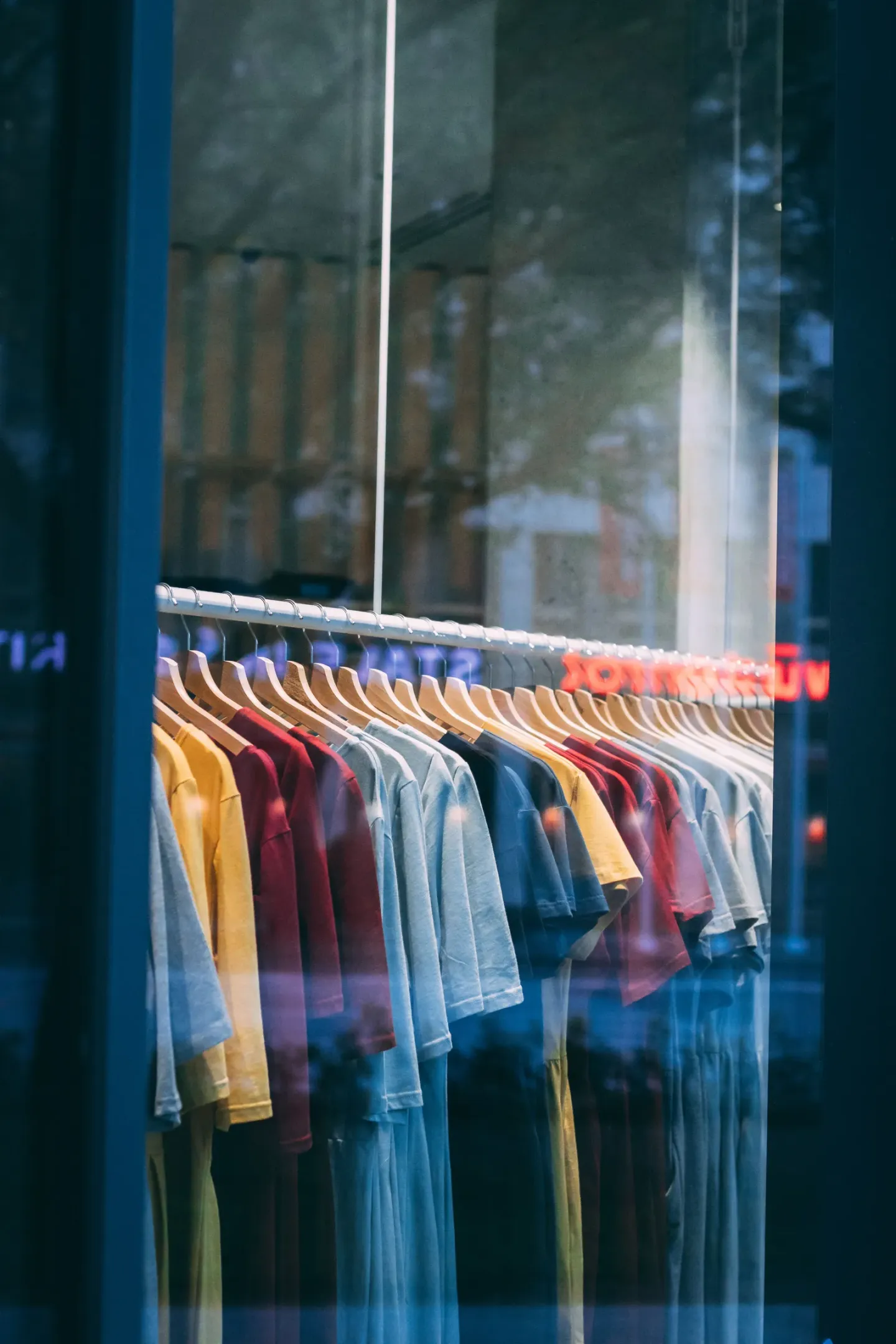 A clothing rail of shirts in muted colours seen through a shop window at dusk, the glass reflecting the street and a neon sign outside.