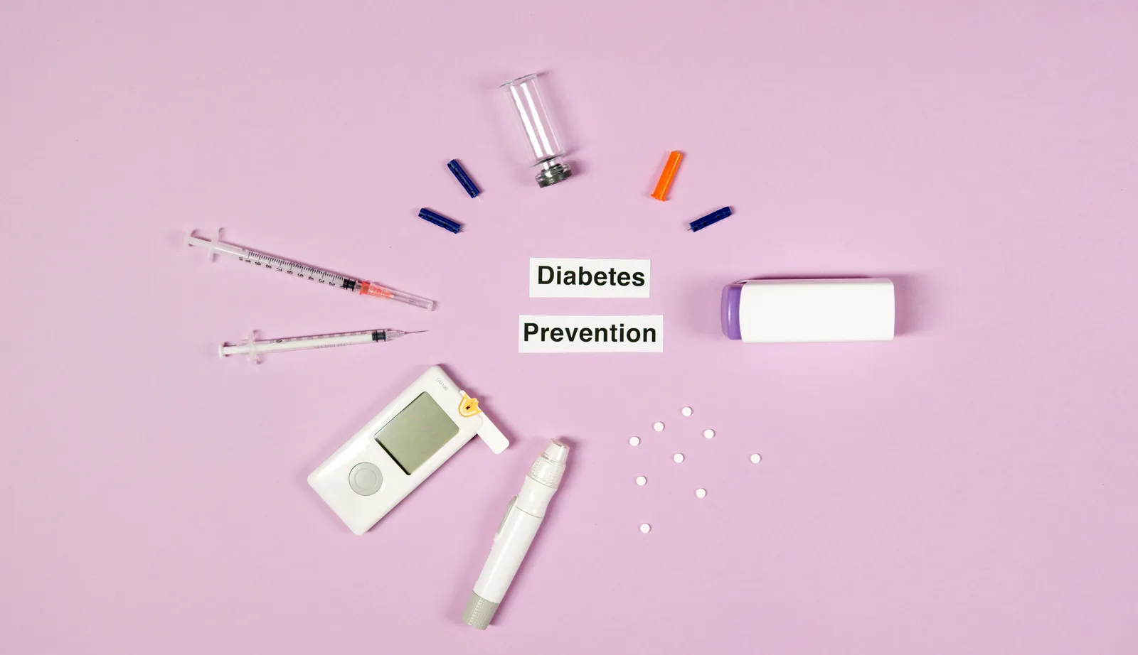 A flat lay on a pink background of diabetes-management supplies — syringes, a glucose meter, a lancing device, vials and loose tablets — arranged around two cards reading Diabetes Prevention.