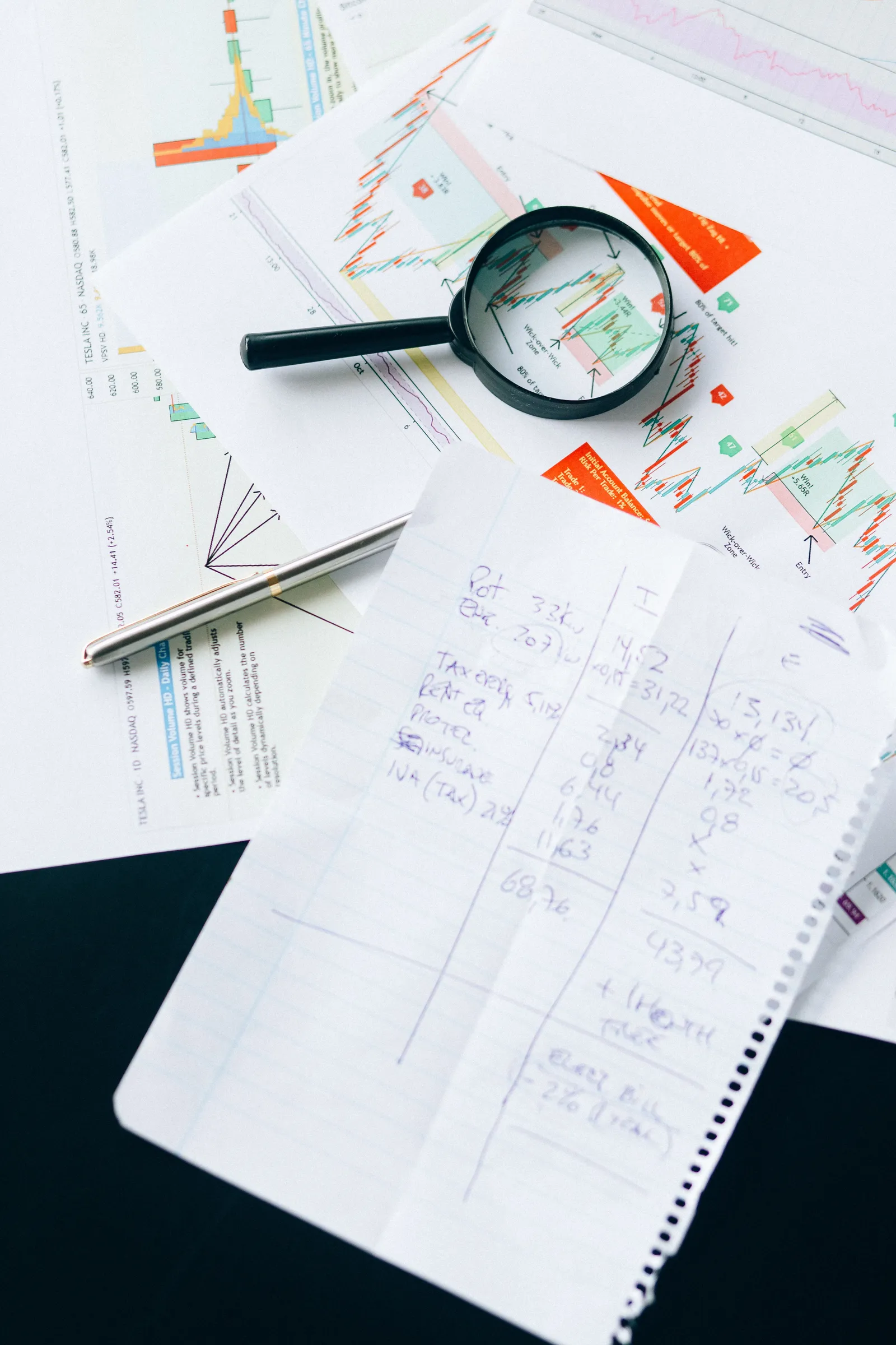 Printed financial charts and handwritten calculations spread across a desk with a pen and a magnifying glass resting on top of a line graph.