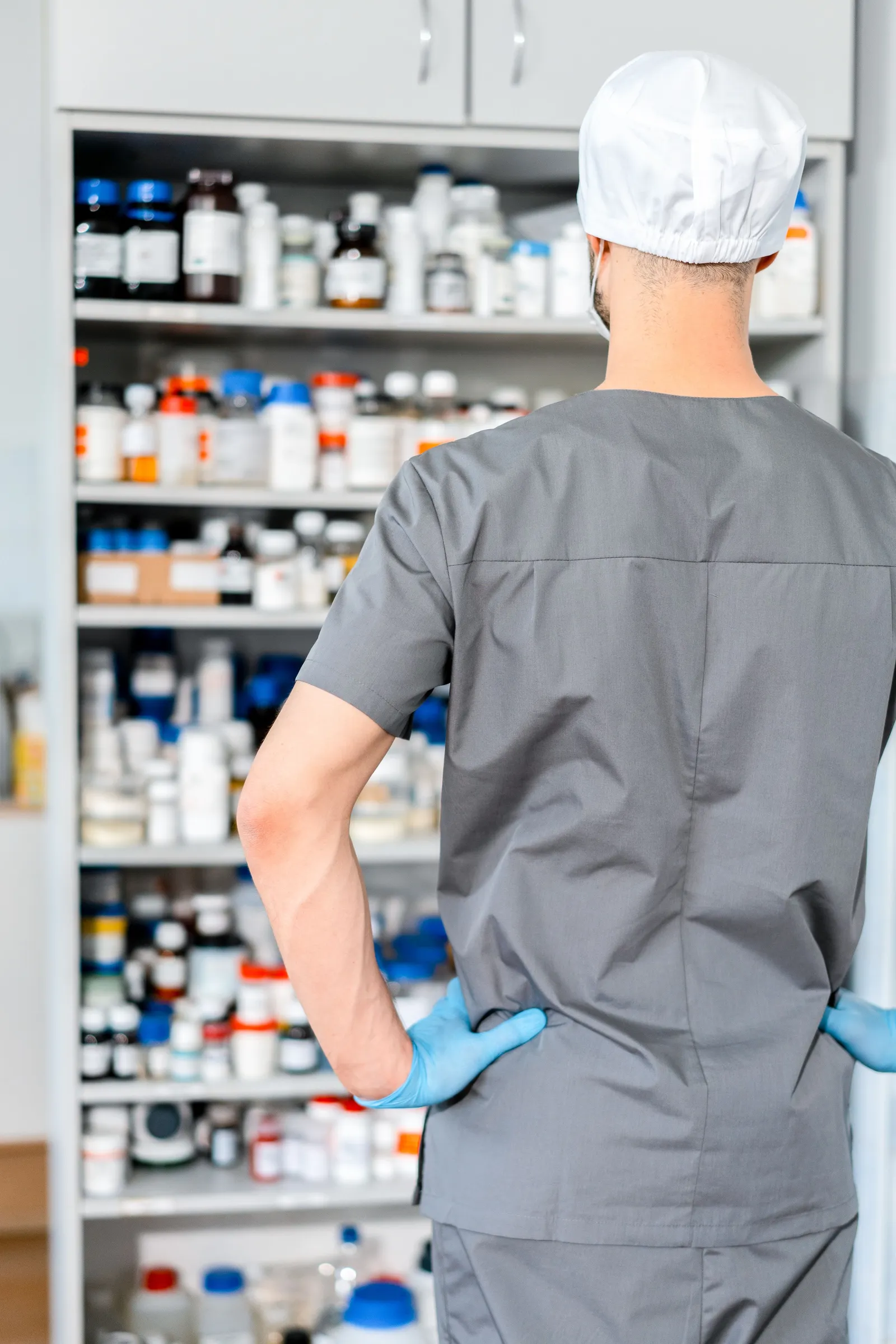 A pharmacist in scrubs, a surgical cap and blue gloves standing with their back to the camera, facing floor-to-ceiling shelves of stock bottles and packets.