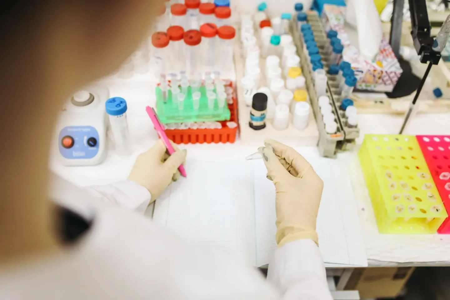 A gloved technician writing notes at a laboratory bench crowded with racks of capped sample tubes, pipettes and a yellow reagent tray.