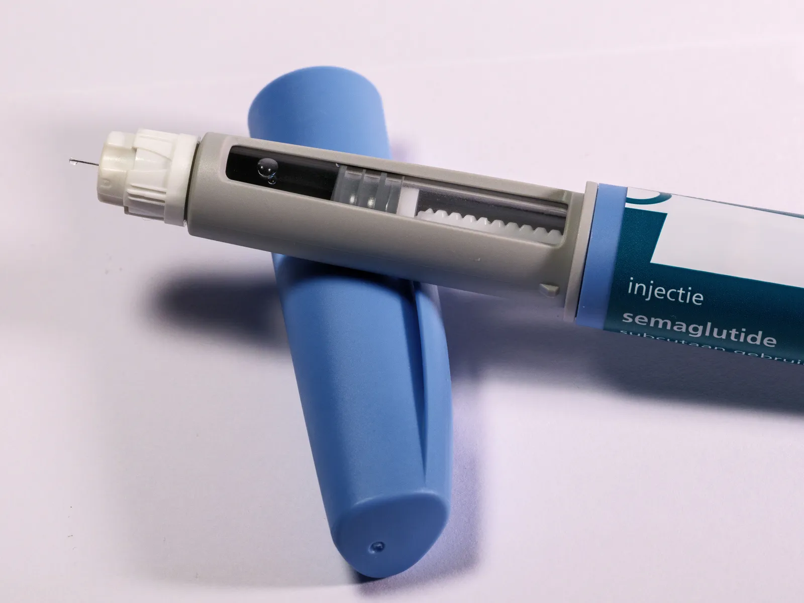 A close-up of a semaglutide injection pen lying on a pale surface beside its blue cap, the dose window and plunger visible along the barrel and the drug name printed on the side.