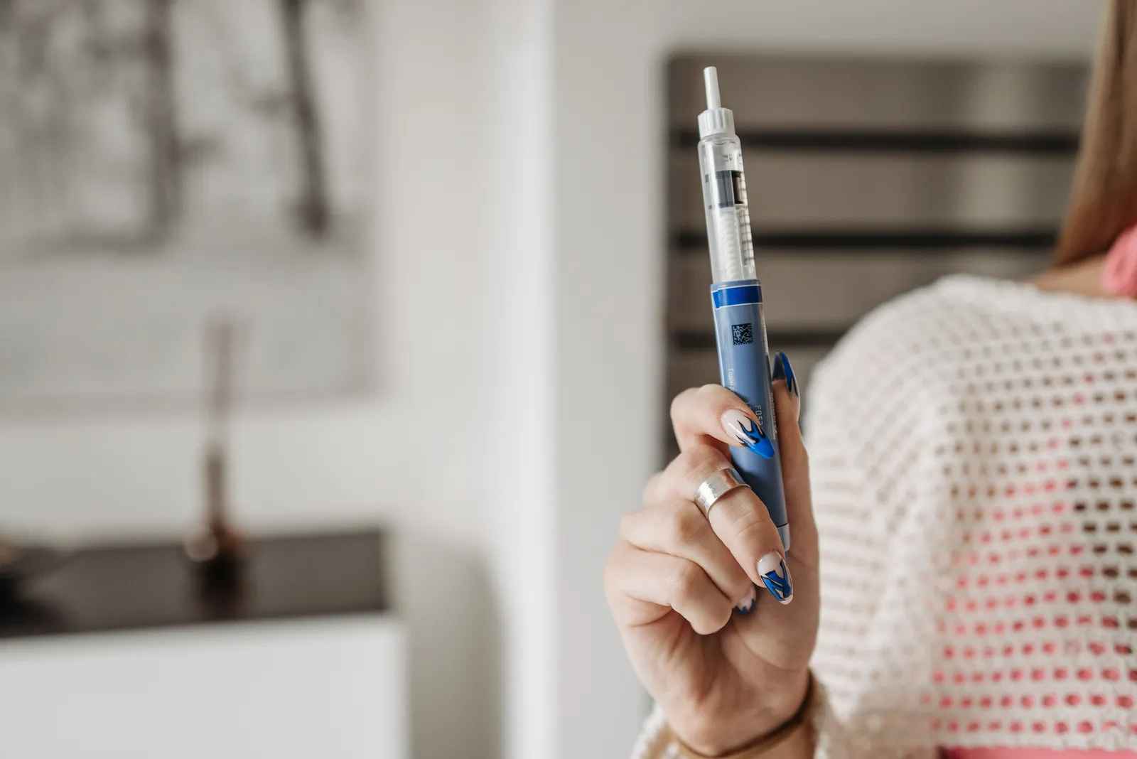 A woman holding up an injection pen and dialling the dose, photographed from behind her shoulder in a softly lit room.