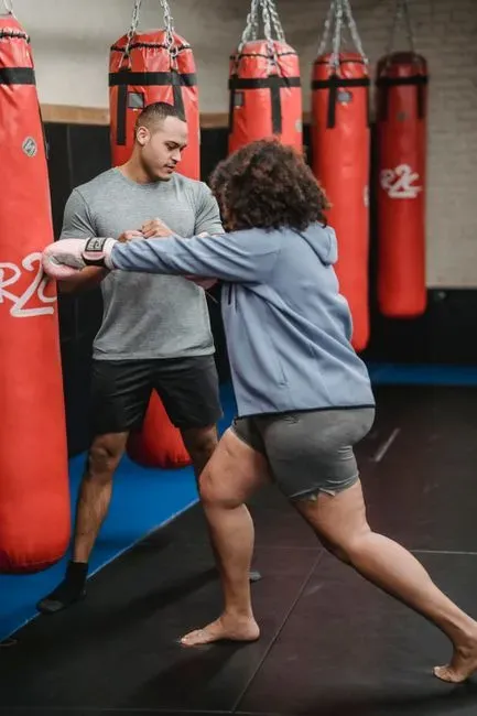 A trainer coaching a plus-size woman through a strike drill in a boxing gym, red heavy bags hanging in a row behind them.