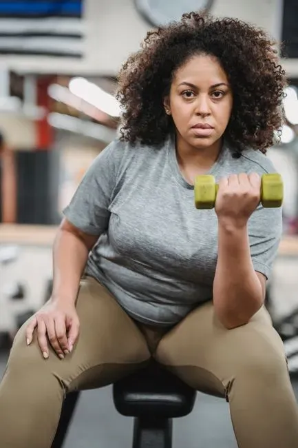 A plus-size woman seated on a workout bench mid bicep curl with a green dumbbell, looking straight at the camera.